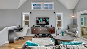 A modern living room with a high ceiling, a flat-screen TV, a dark wooden entertainment center, a grey armchair, a round coffee table, and a large window with a view.