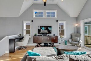 Een moderne woonkamer met een hoge nok, een flatscreen televisie, een donker houten tv-meubel, een grijze fauteuil, een ronde salontafel en een groot raam met uitzicht.