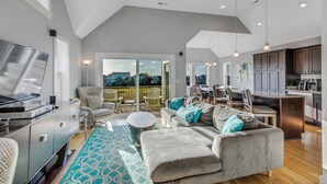 A spacious living area with a gray sofa, a coffee table, and a TV. There is a balcony with a view of the outdoors.