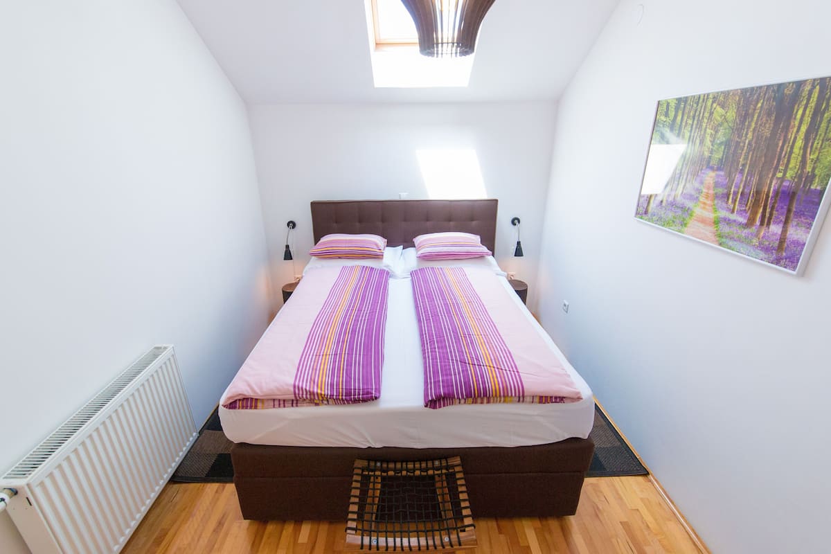 Een slaapkamer met een groot bed, een radiator, een dakraam en een schilderij met een boslandschap.
