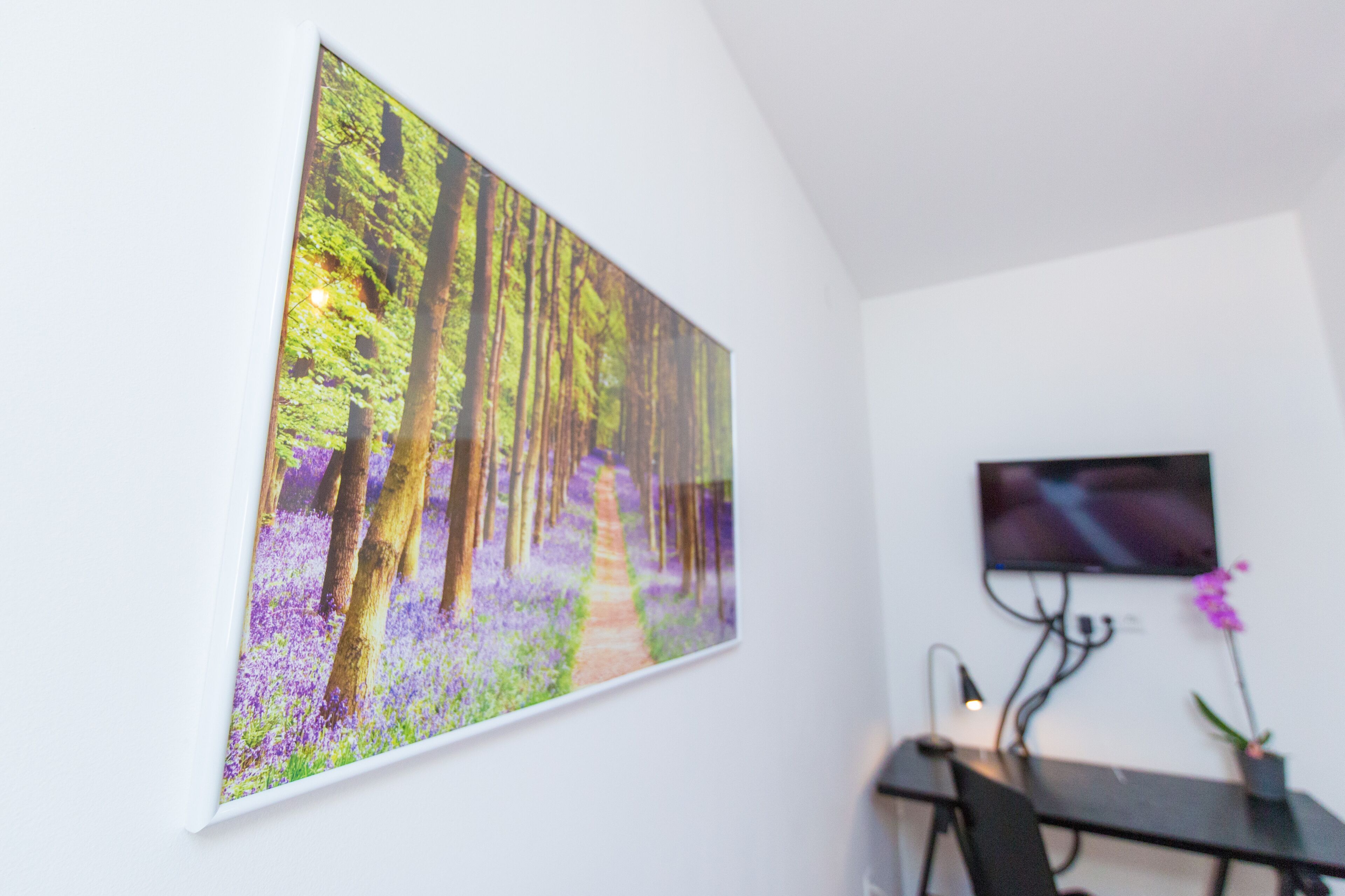 A room with a large forest-themed painting, a flat-screen TV mounted on the wall, and a desk with a chair.