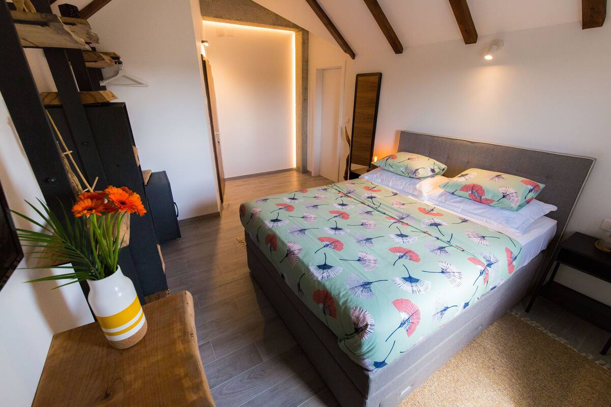 Een slaapkamer met een bed, nachtkastjes, een vloer van hout, een vaas met bloemen en een deur naar een andere kamer.