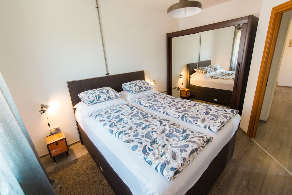Een tweepersoonsbed met beddengoed met een patroon, een nachtkastje met een lamp en een grote spiegel boven het bed.