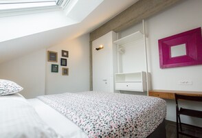 Un dormitorio con tragaluz, una cama con colcha floral, un armario blanco, un escritorio de madera y un espejo con marco rosa.