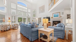 A spacious living room with a high ceiling, large windows, and a fireplace.