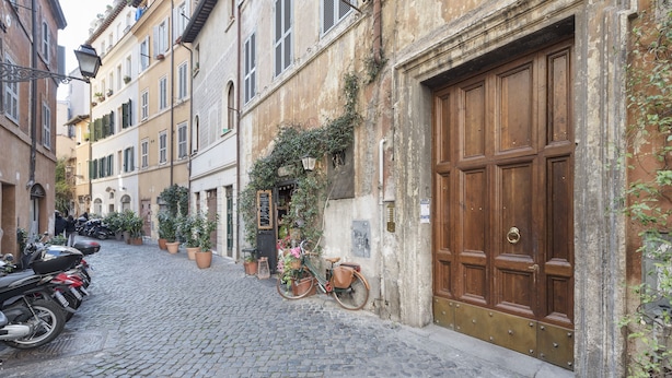 A cobblestone street with parked motorcycles and bicycles, a large wooden door, and buildings with balconies and potted plants.