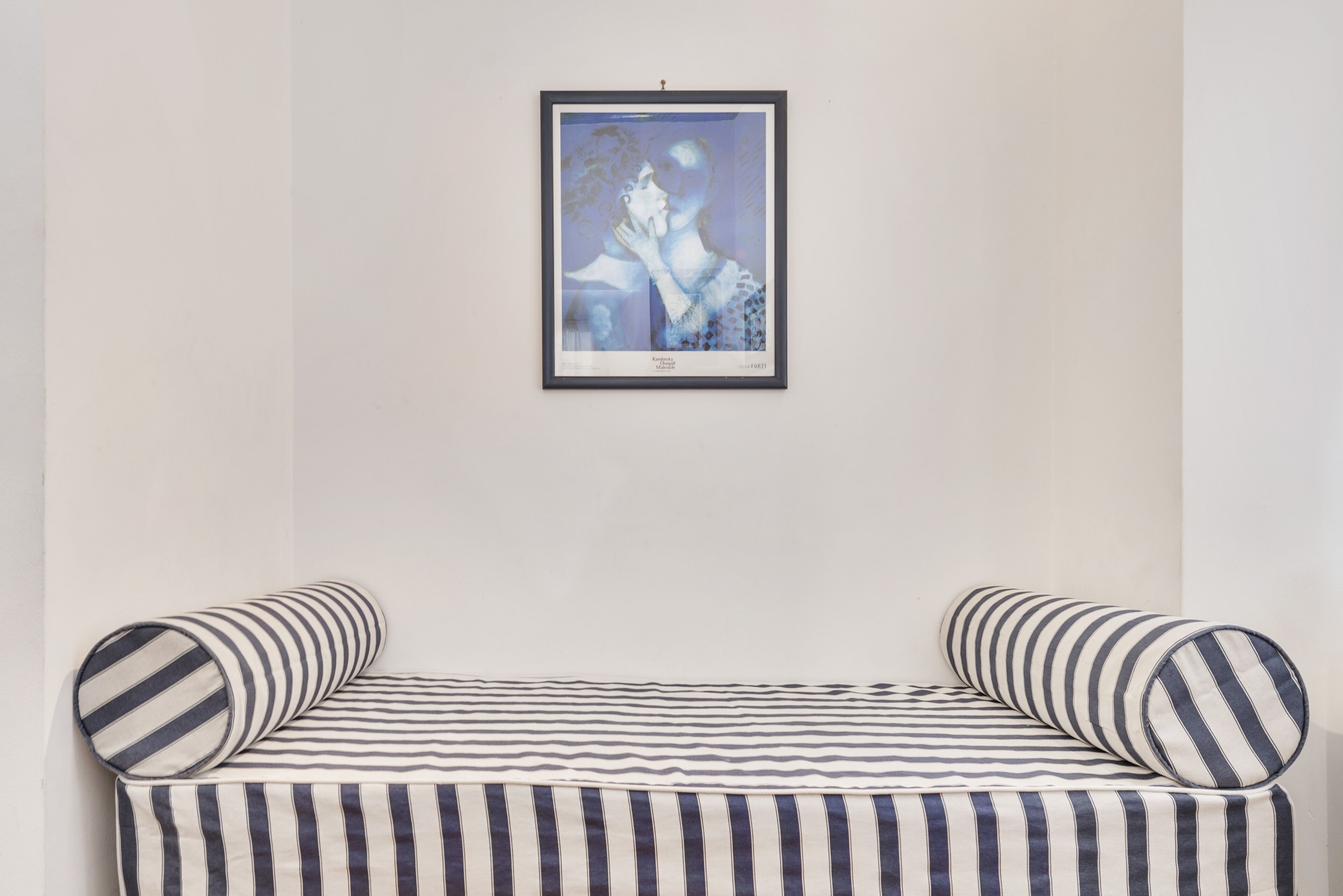 A bed with striped bedding and pillows, a framed artwork above it, and a plain wall.