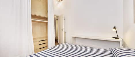 A bedroom with a bed, a wooden wardrobe, a white bedside table, and a hanging light.