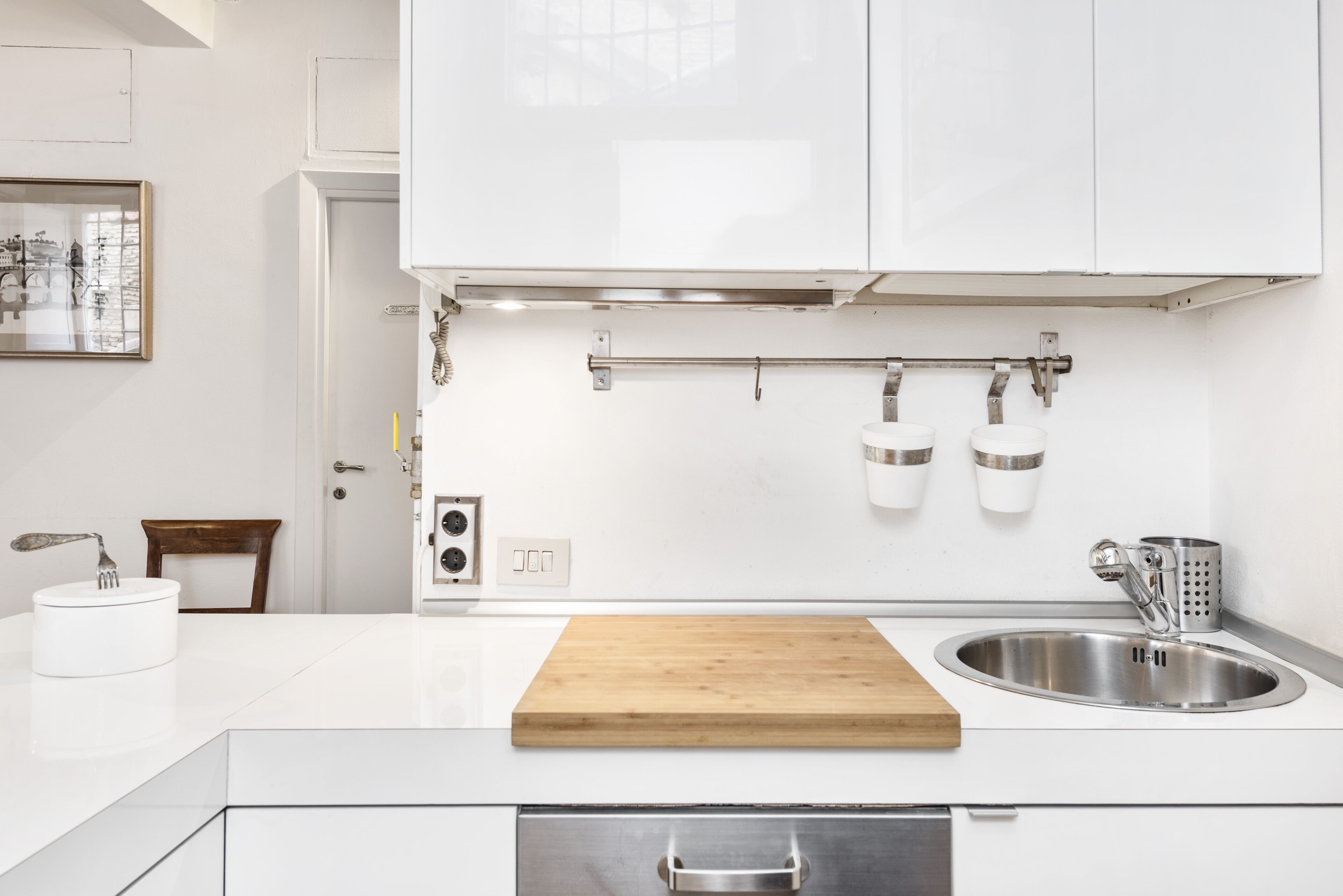 Una cocina moderna con armarios blancos, una tabla de cortar de madera, un lavamanos de acero inoxidable y un colgador para ollas.