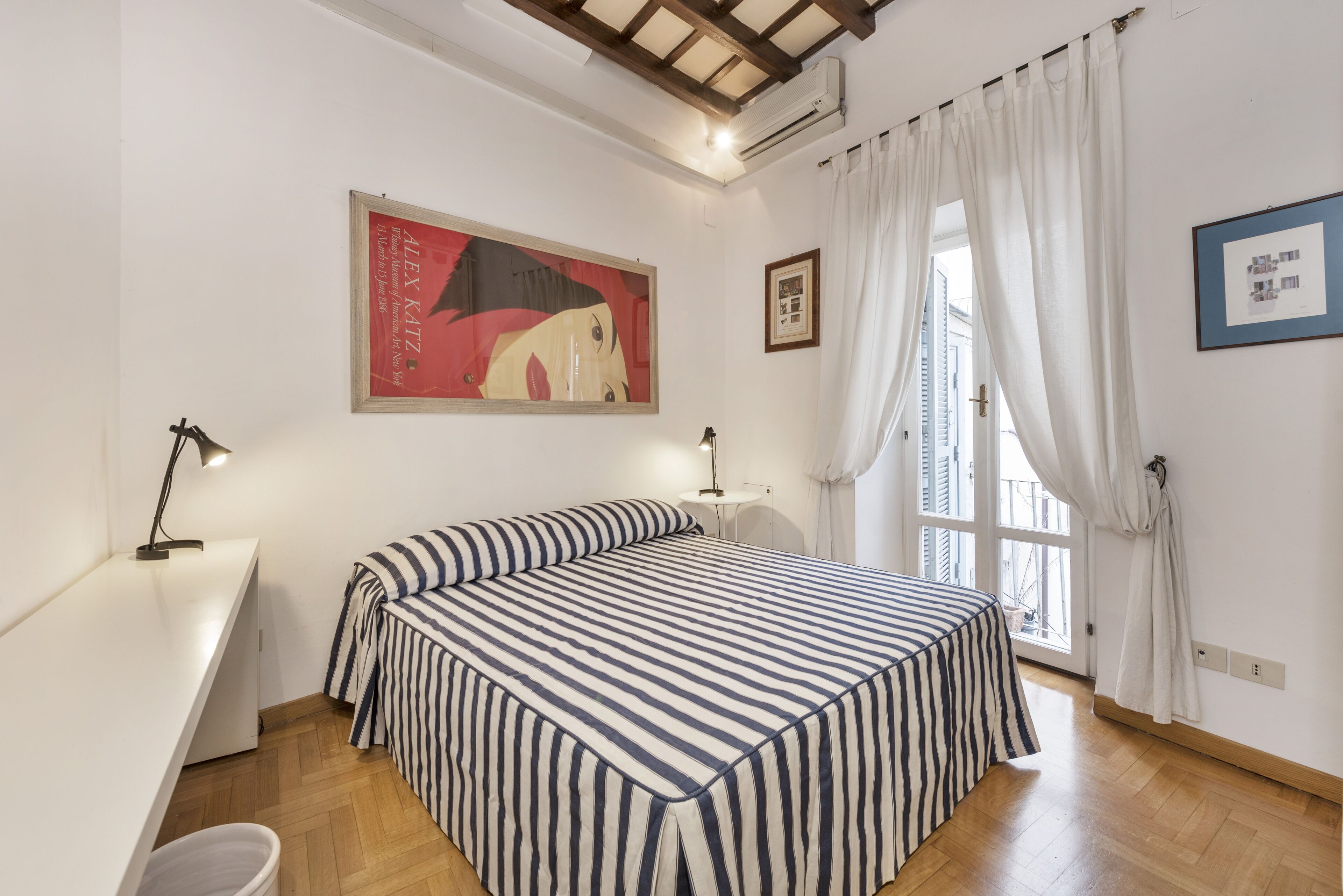A bedroom with a bed, a desk, a chair, a window with curtains, and a painting on the wall.