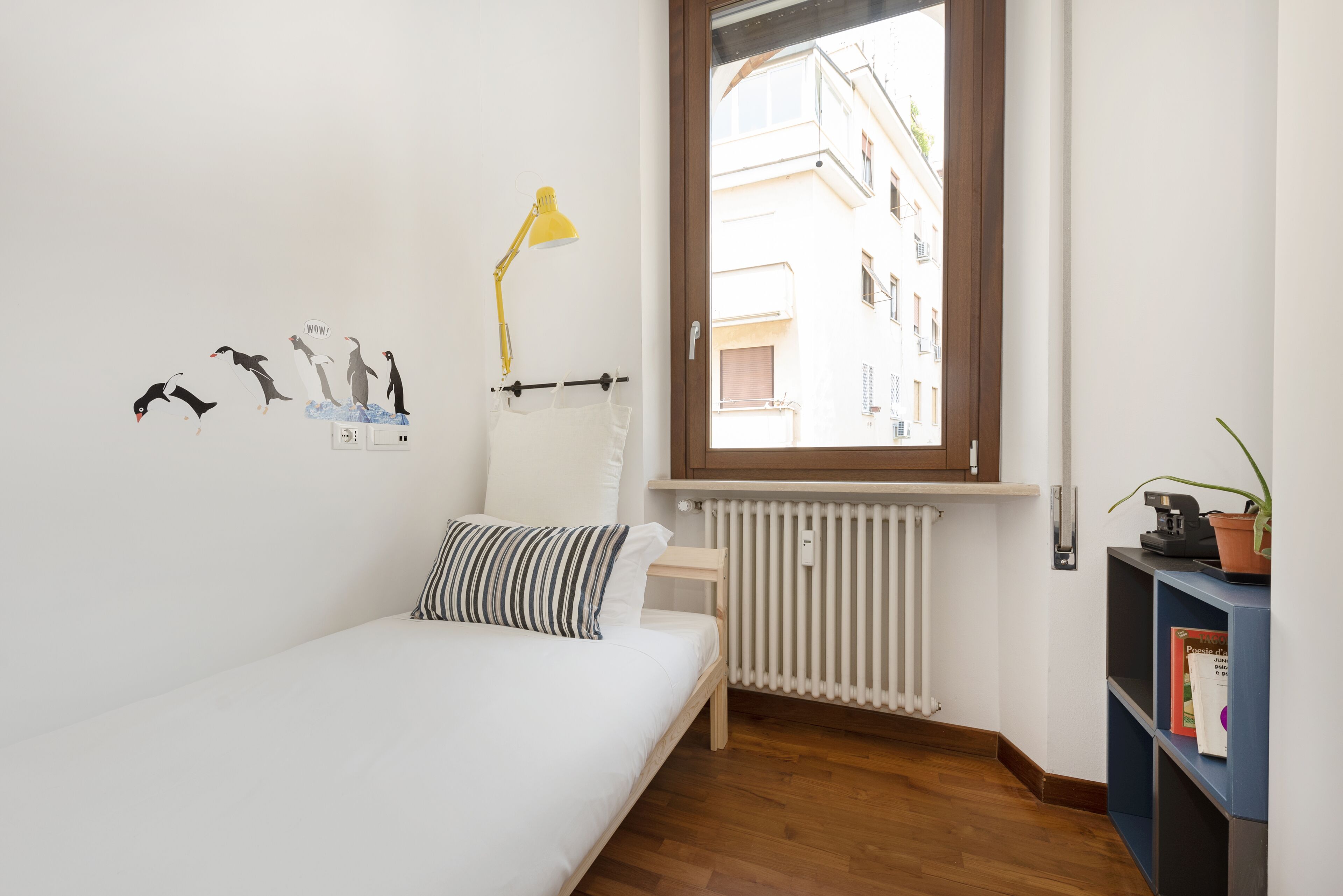 A bedroom with a bed, a window, a radiator, a lamp, and a bookshelf.