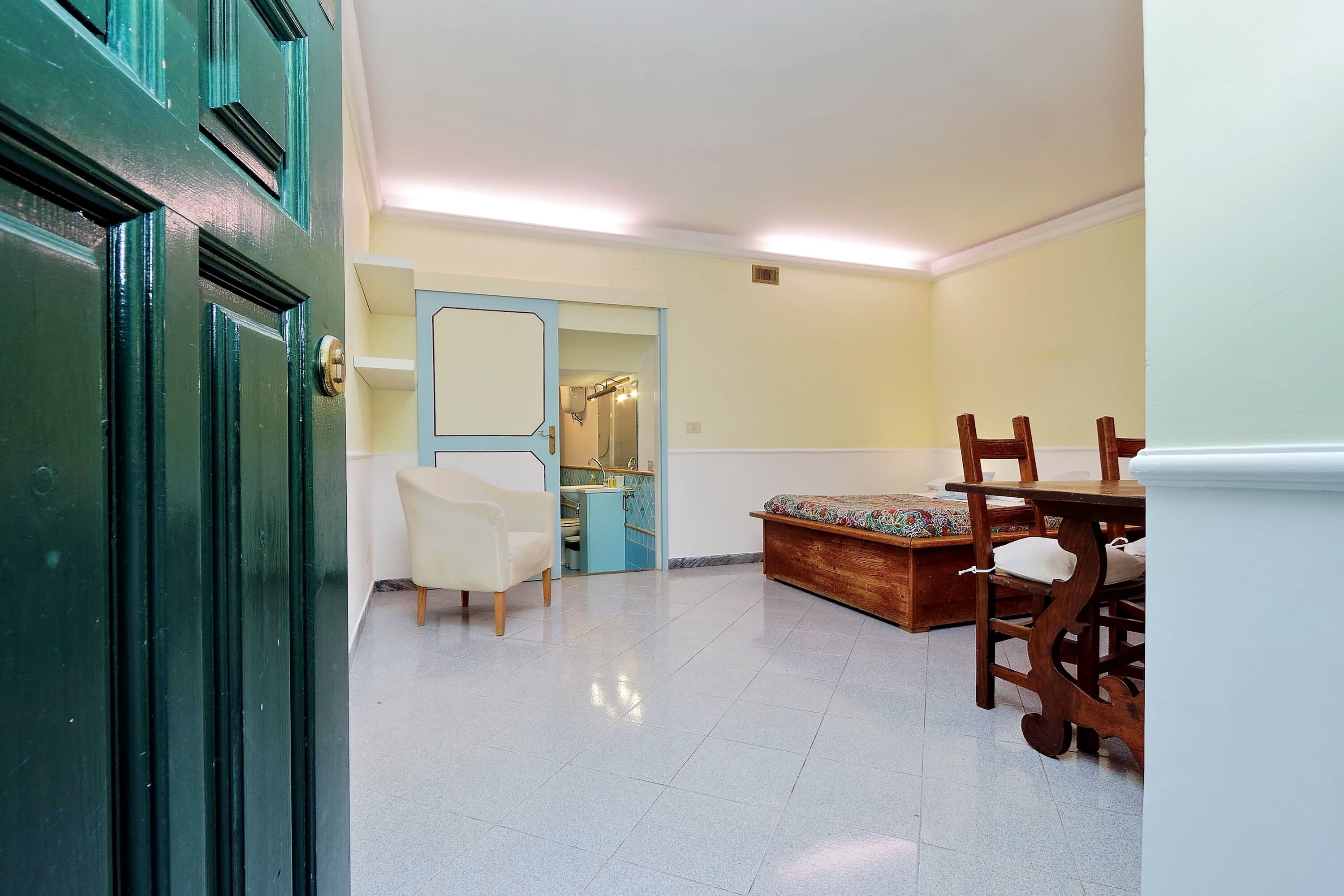 A room with a bed, a chair, a small table, a mirror, and a door.