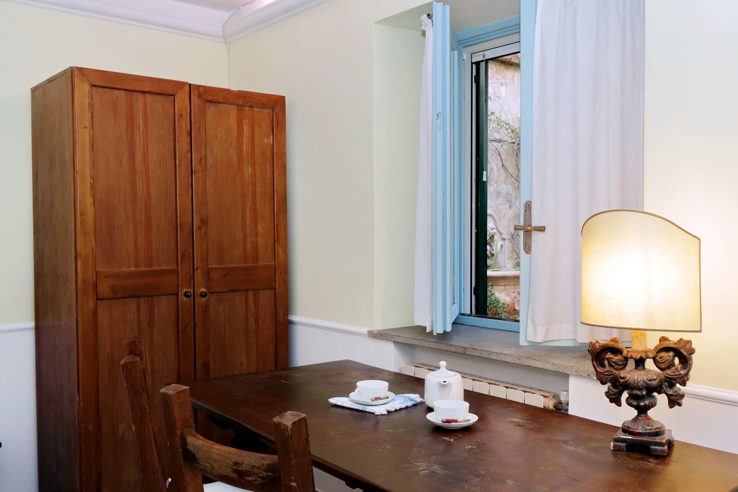 Une table de salle à manger en bois, une théière blanche et des tasses, une fenêtre avec des volets bleus et une armoire en bois.