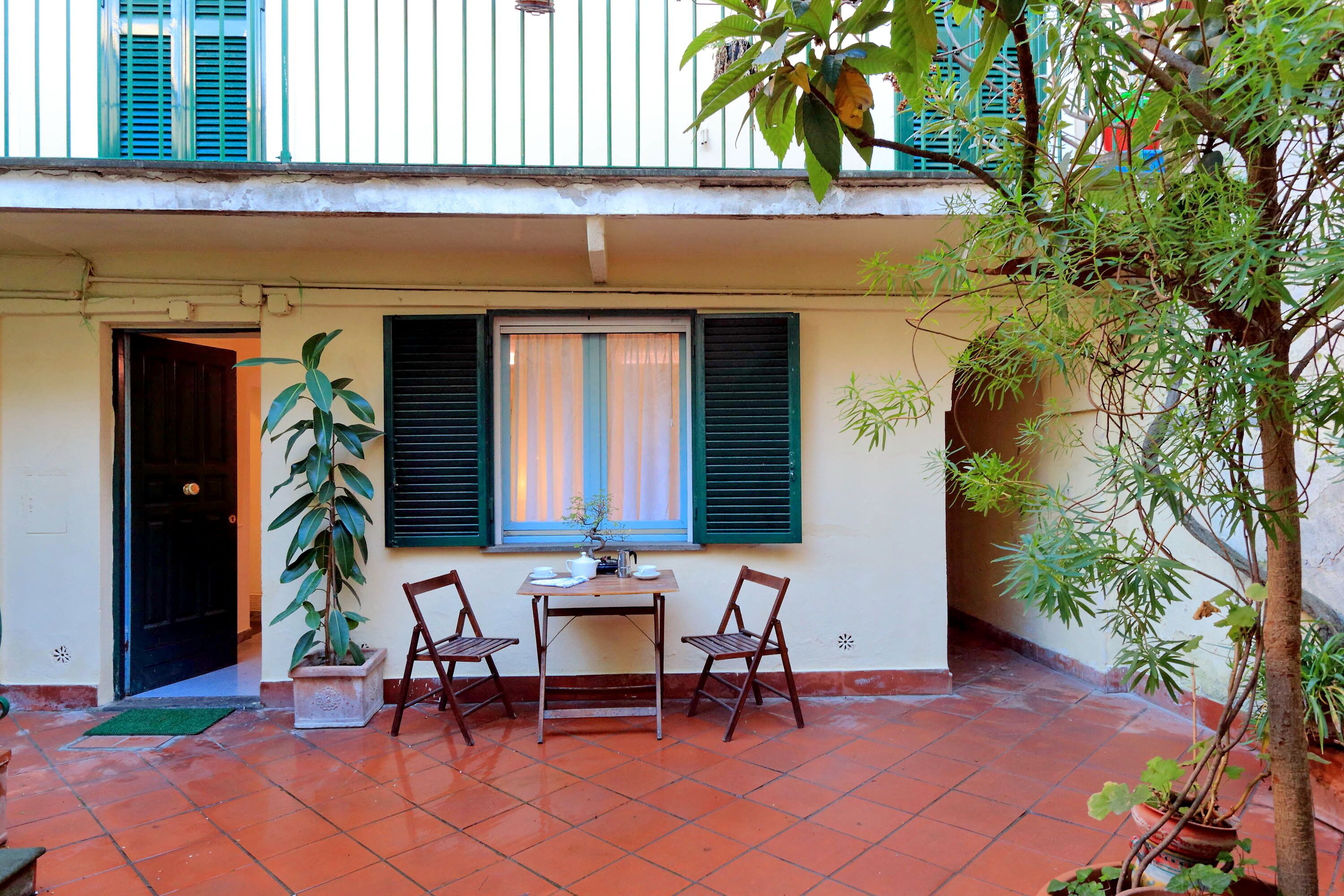 Eine kleine Terrasse im Freien mit Tisch und Stühlen, einem Blumentopf und einem Fenster mit Rollläden.