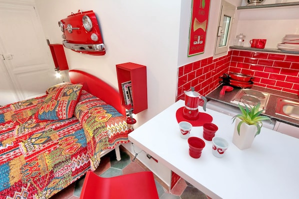 A room with a bed, a white table with red cups, a red chair, a red shelf, and a red wall decoration.