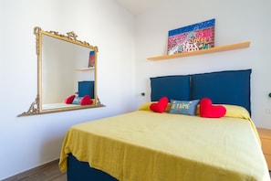 Ein ordentlich bezogenes Bett mit gelber Tagesdecke und blauem Kopfteil, ein großer Spiegel mit aufwendigem Rahmen sowie ein farbenfrohes Graffiti-Kunstwerk an der Wand.