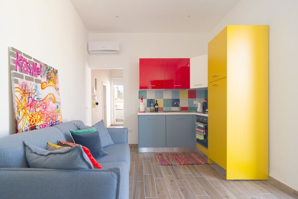Ein modernes Wohnzimmer mit einem grauen Sofa, bunten Kissen und einem lebhaften abstrakten Gemälde. Im Hintergrund ist eine Küche mit roten und gelben Schränken zu sehen.
