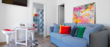 A modern living room with a grey sofa, red and green cushions, a white coffee table, and two red bar stools. A colorful graffiti artwork is on the wall.