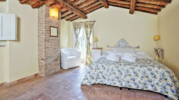 A bedroom with a stone wall, a wooden ceiling, a bed with a floral bedspread, a white chair, and a nightstand with a lamp. - Volpe (Rome)
