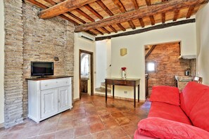 A living room with a brick wall, a wooden ceiling, a red sofa, a TV on a white cabinet, and a staircase leading to another room. - Volpe (Rome)