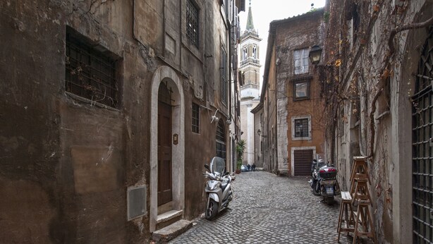 A narrow cobblestone street with historic buildings, a church tower, and parked scooters. - Volpe grande (Rome)