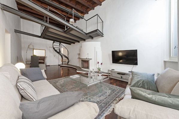 A modern living room with a glass coffee table, beige sofas, a staircase, and a flat-screen TV.