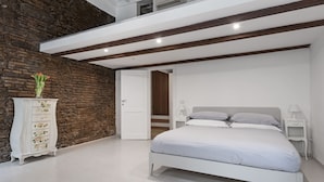 A bedroom with a white bed, a dresser with floral patterns, a potted plant, and a stone wall. - Volpe grande (Rome)