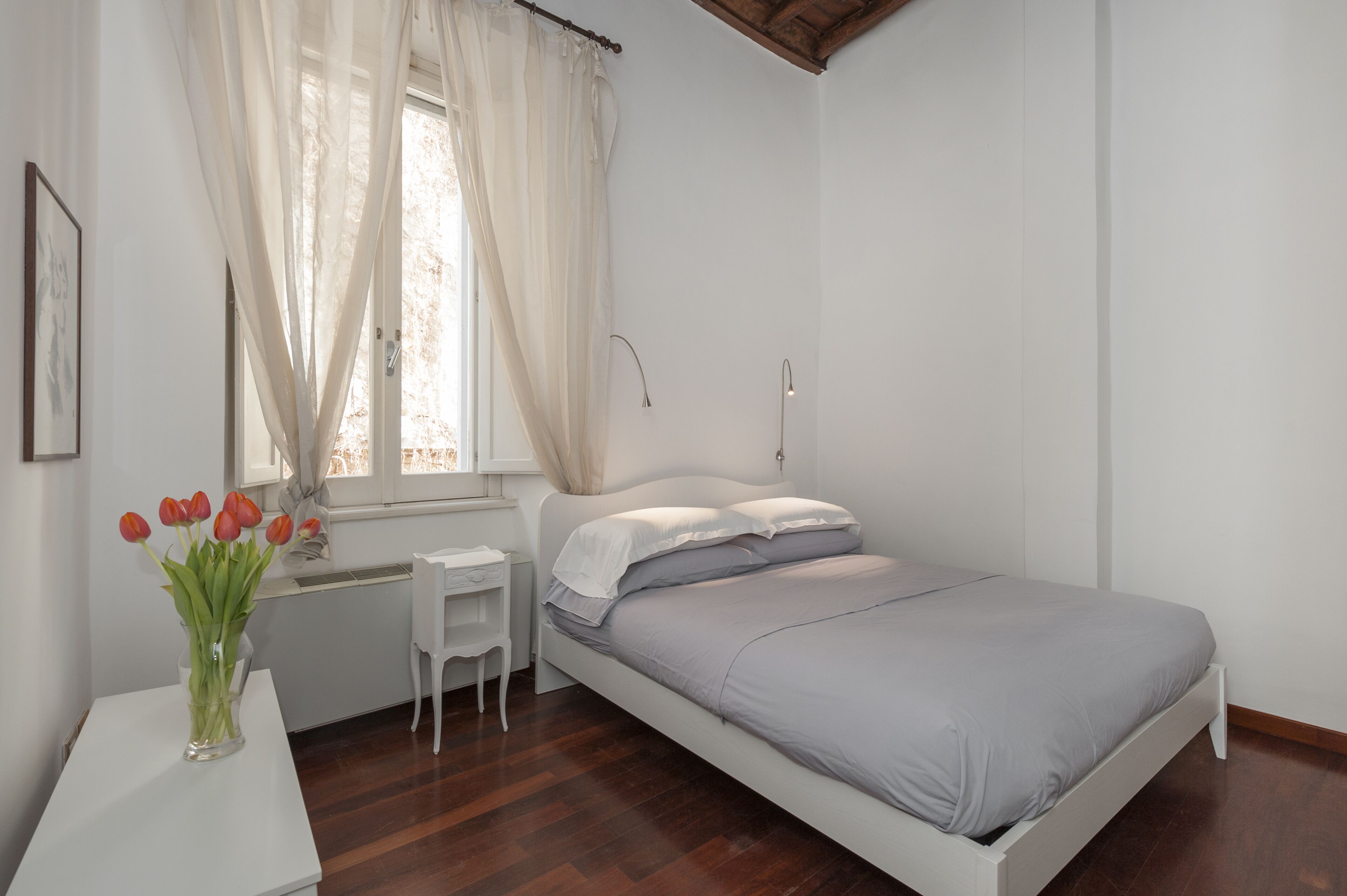 A bedroom with a bed, a bedside table, a window with curtains, and a vase with flowers.