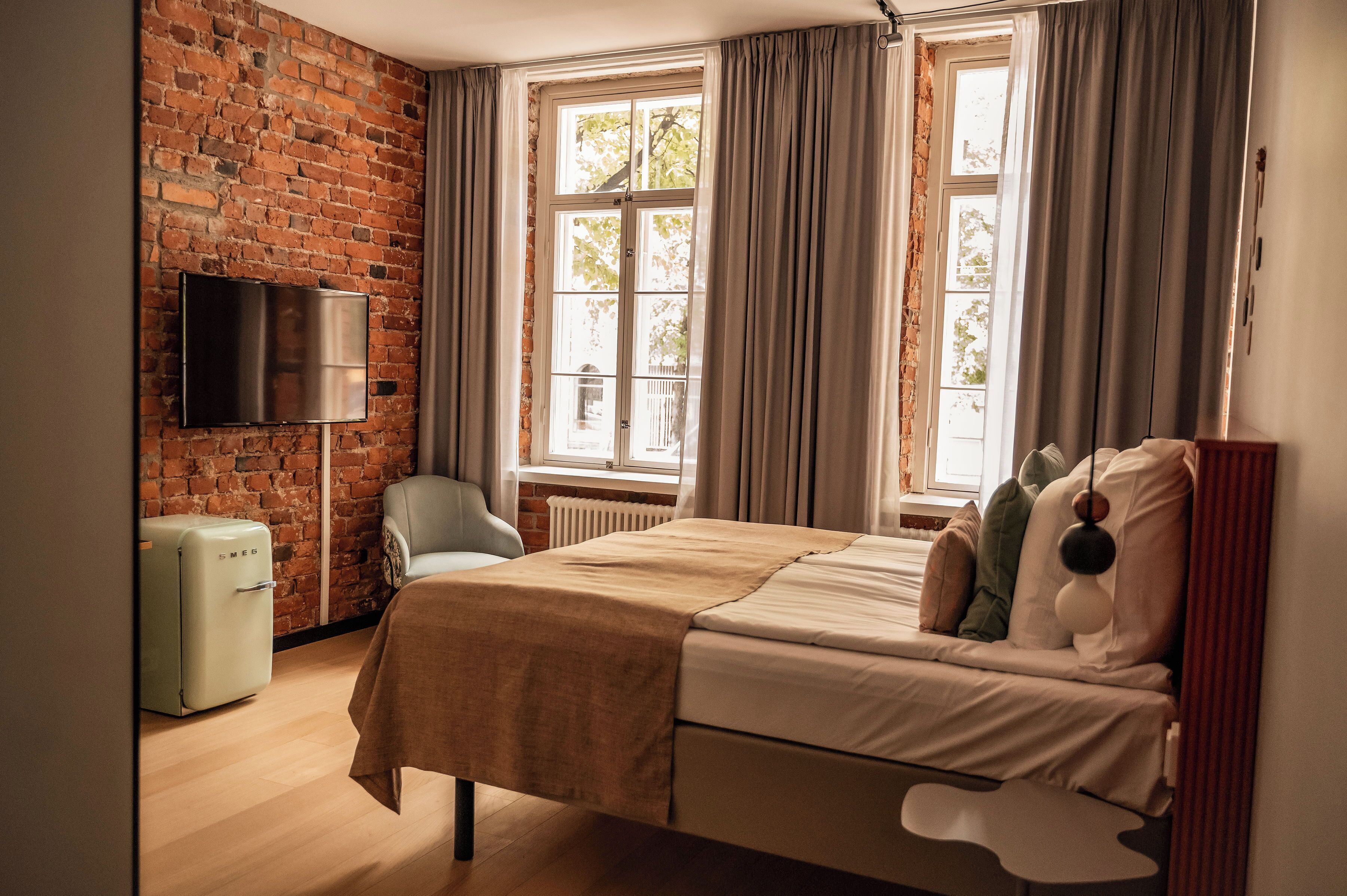 Una habitación con pared de ladrillo, cama, televisor, silla y ventana con cortinas.