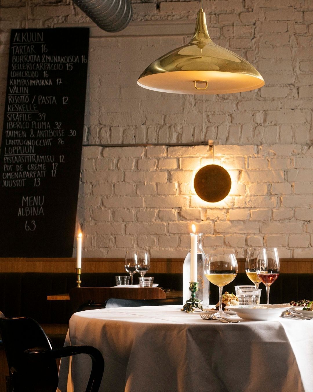 A restaurant interior with a brick wall, a hanging pendant light, a menu on the wall, and a table set for dining.