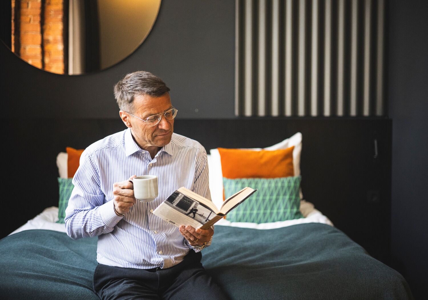 A man sitting on a bed with a book and a mug, in a room with a circular mirror and striped pillows.