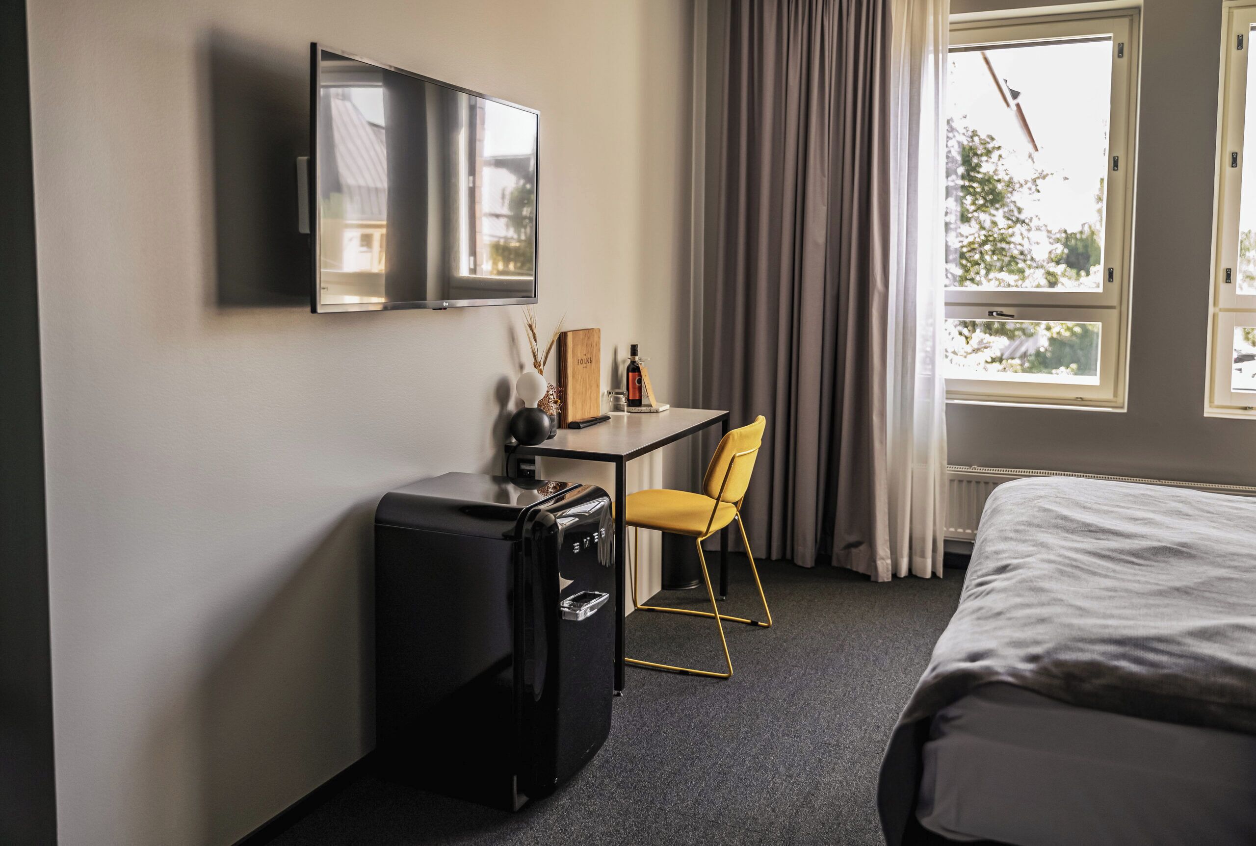 Habitación de hotel con cama, escritorio con silla, televisor montado en la pared y ventana con cortinas.