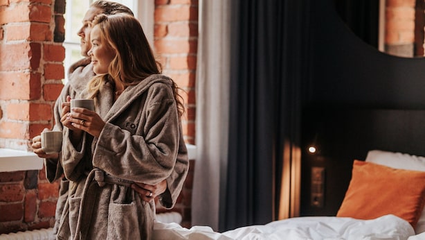 Una habitación con pared de ladrillo, una ventana, una cama con ropa blanca y una persona en bata sosteniendo una taza.