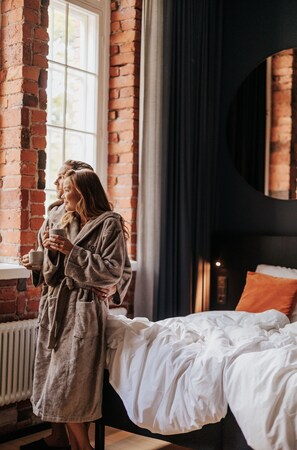 A room with a brick wall, a window, a bed with white bedding, and a person in a robe holding a mug.