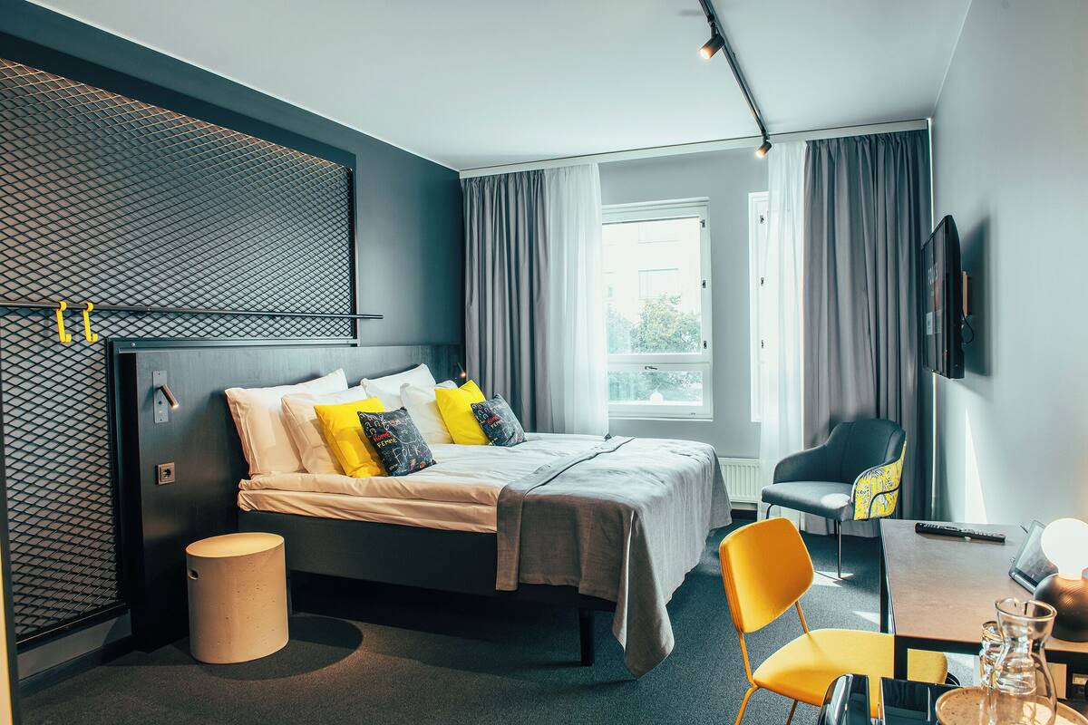 Una habitación de hotel moderna con una cama, un escritorio con silla, un televisor y un ventanal con cortinas.