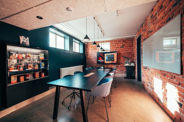 A modern meeting room with a long table, chairs, and a brick wall. There is a whiteboard and a shelf with various items.
