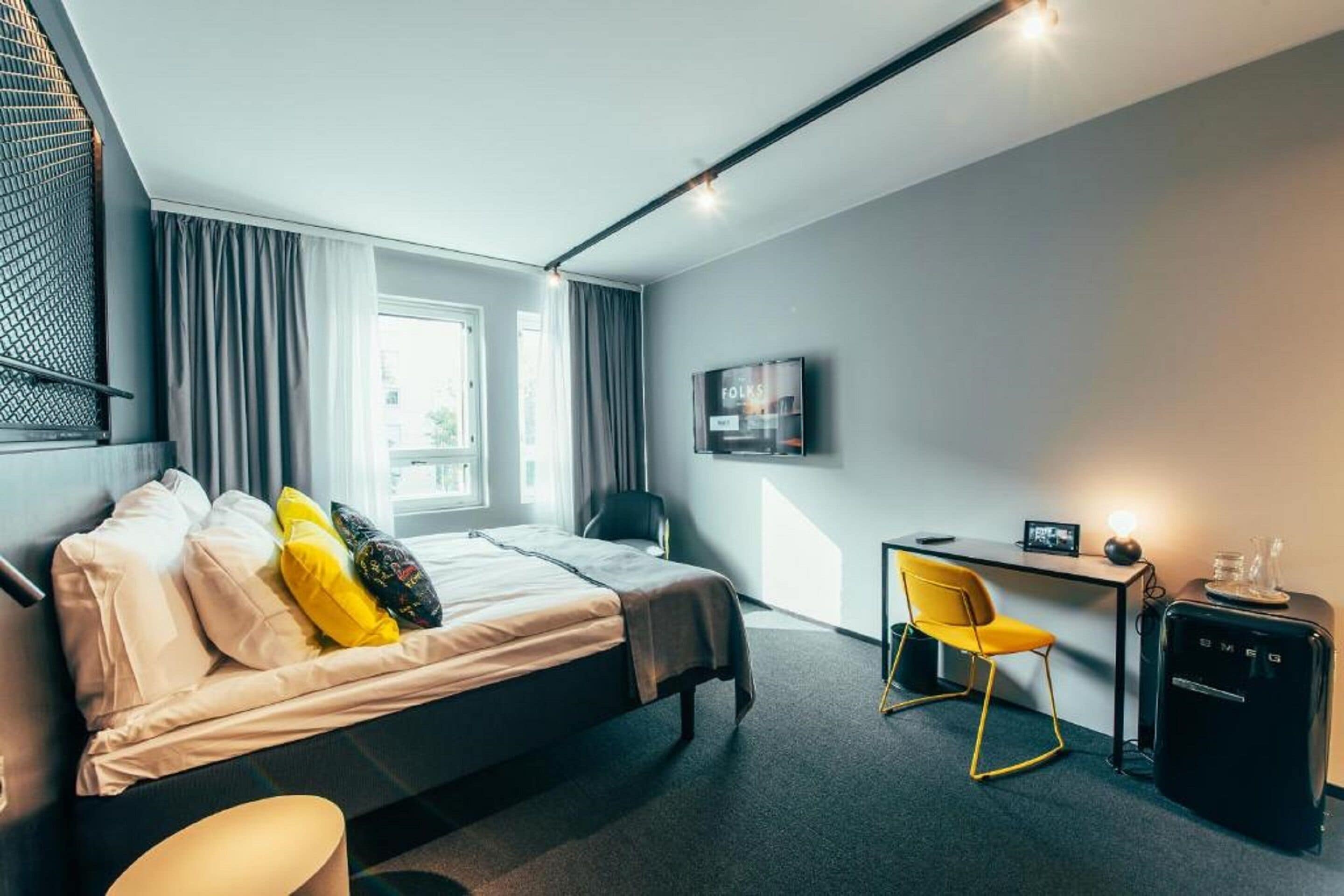 A modern hotel room with a bed, a desk, a chair, a TV, and a minibar.