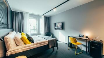 A modern hotel room with a bed, a desk, a chair, a TV, and a minibar.