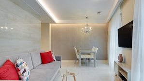 Ein modernes Wohnzimmer mit grauem Sofa, roten und bunten Kissen, einem Glastisch, einem Essbereich mit weißem Tisch und Stühlen, einer Chandelüster und einem Flachbildfernseher auf einer weißen Konsole.