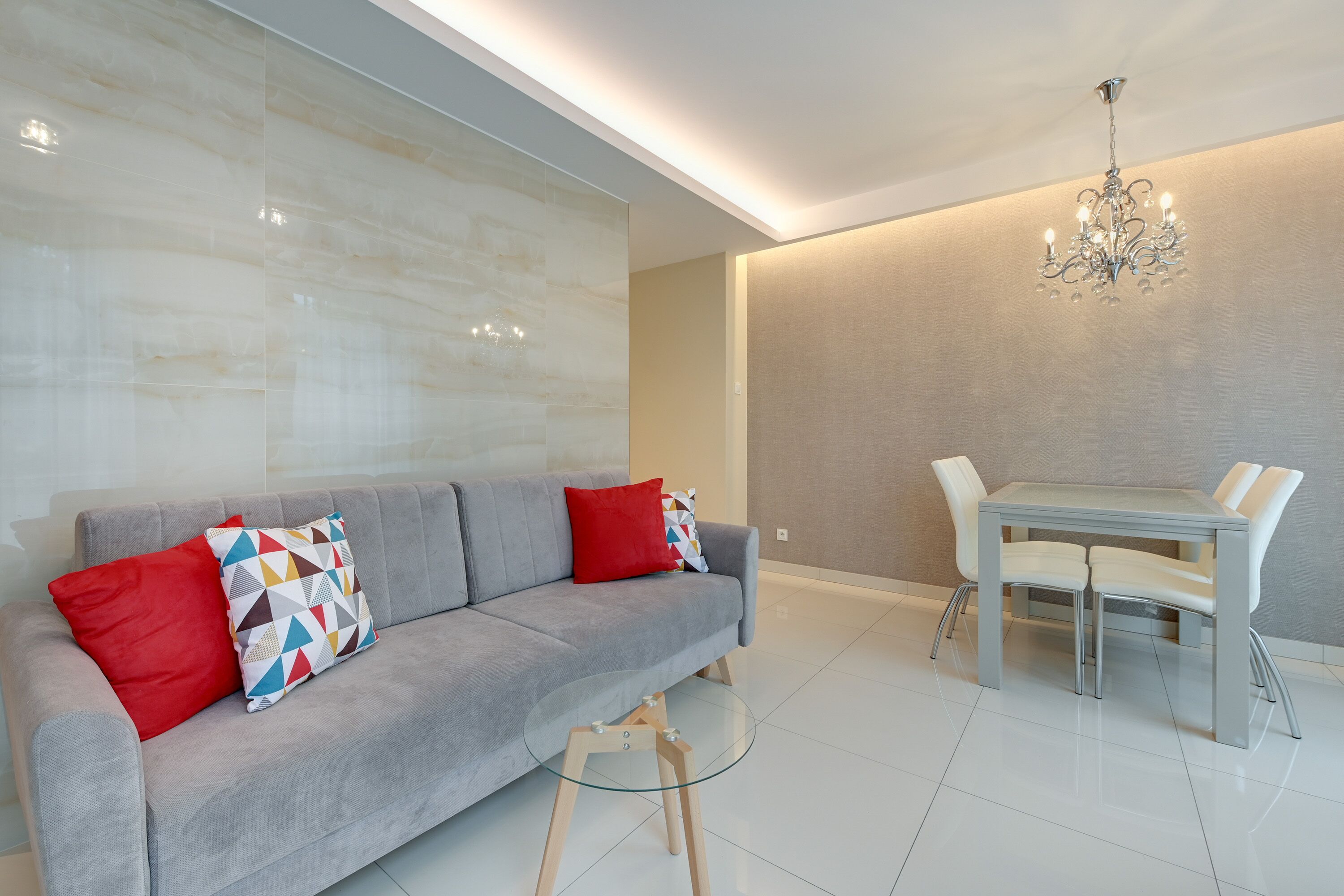 A modern living room with a grey sofa, red and patterned cushions, a glass coffee table, and a dining area with a white table and chairs.