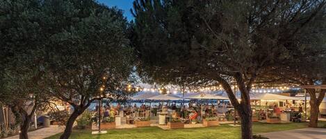 An outdoor dining area with tables and chairs, surrounded by trees and under string lights.