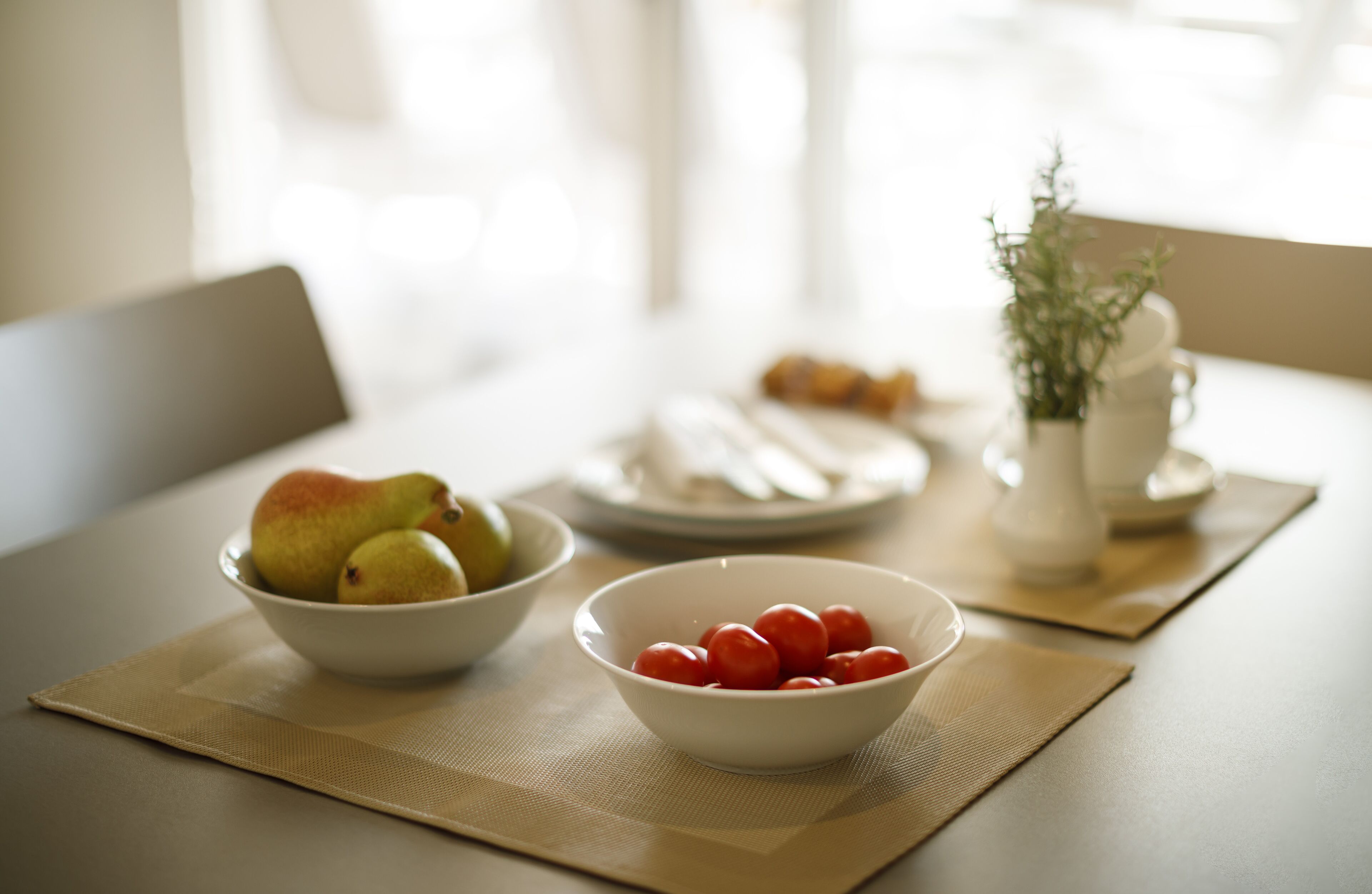 A dining table with a bowl of pears and a bowl of cherry tomatoes, a plate with pastries, and a vase with a plant.