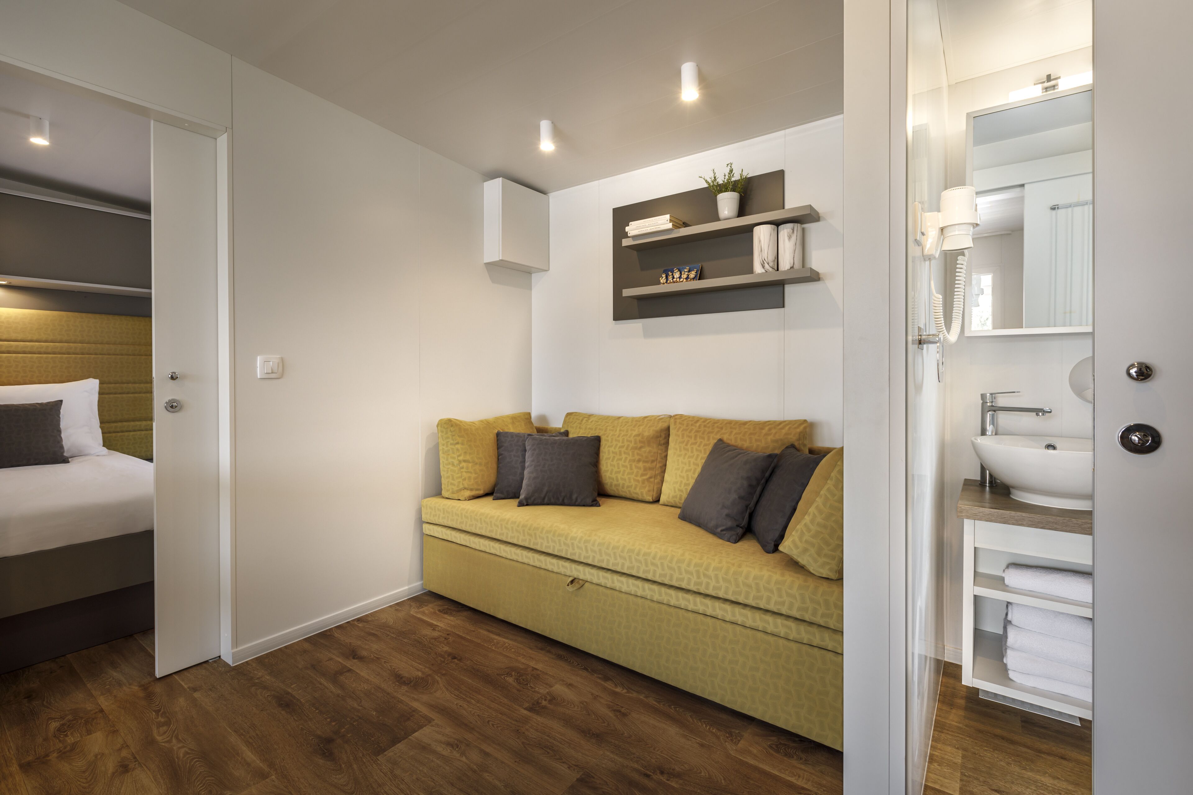 Una habitación de hotel moderna con un sofá amarillo, una cama con cabecera de madera, un estante pequeño con artículos de aseo y un baño con lavamanos y espejo.
