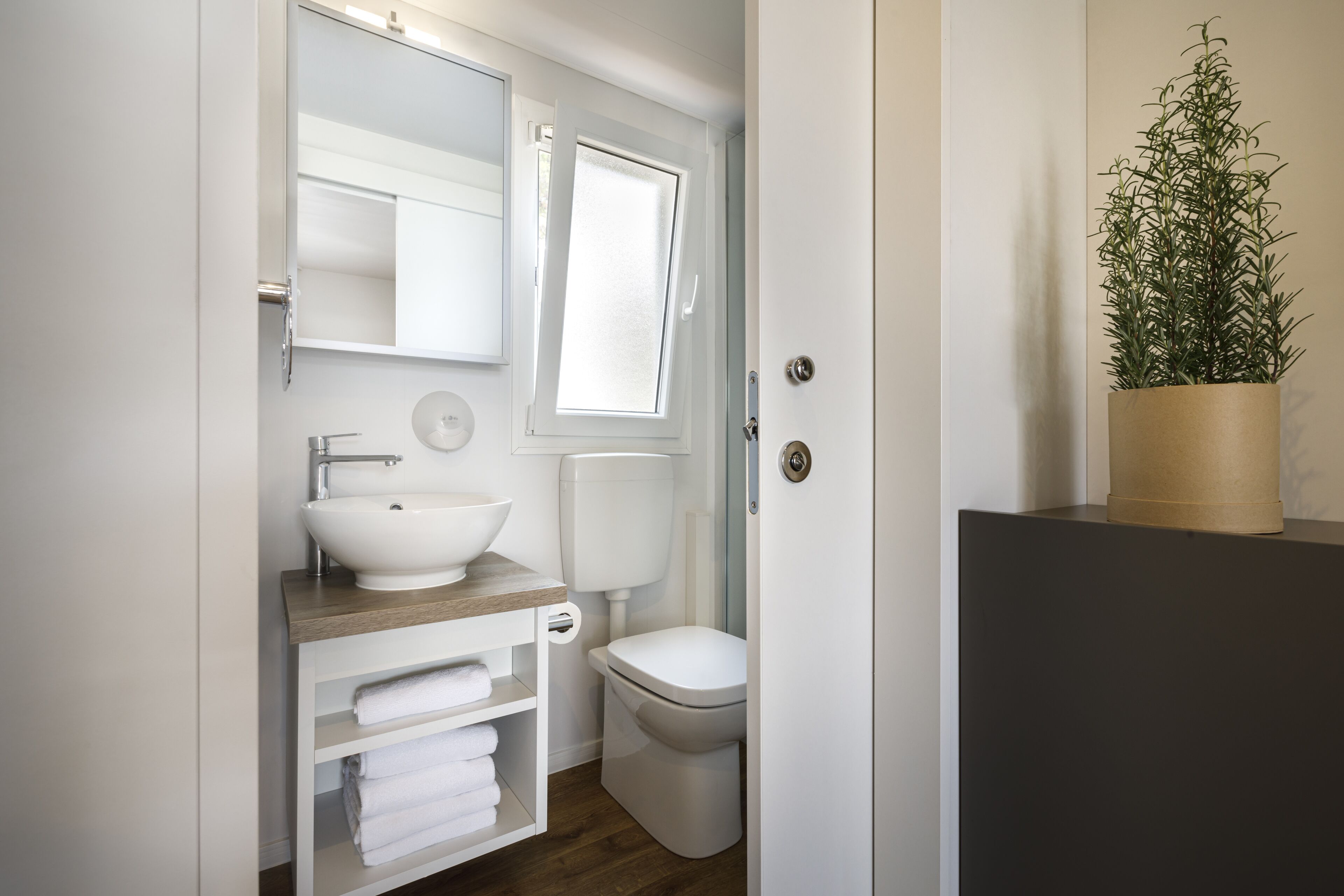 A bathroom with a white sink, toilet, and a mirror above the sink. There is a window above the toilet and a potted plant on a shelf.