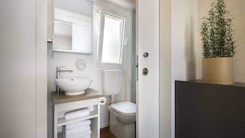 Un baño con un lavamanos blanco, inodoro y un espejo sobre el lavamanos. Hay una ventana sobre el inodoro y una planta en maceta en un estante.