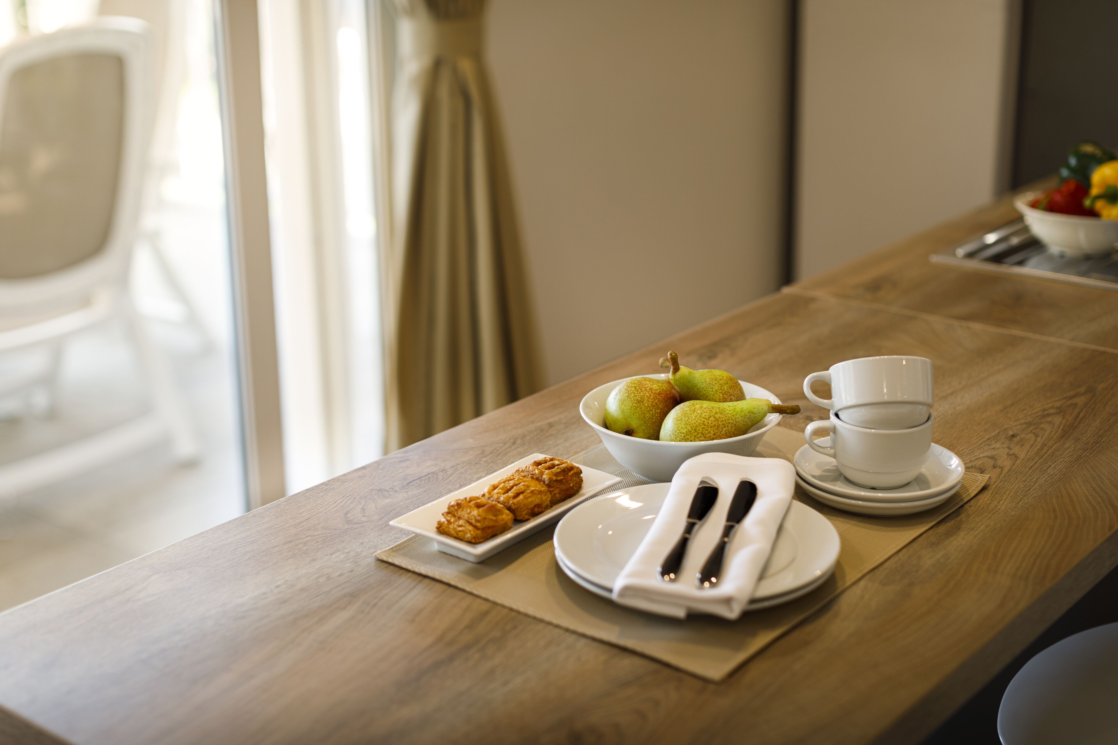 A breakfast table with a bowl of fruit, a plate of pastries, a cup, and a napkin with cutlery.