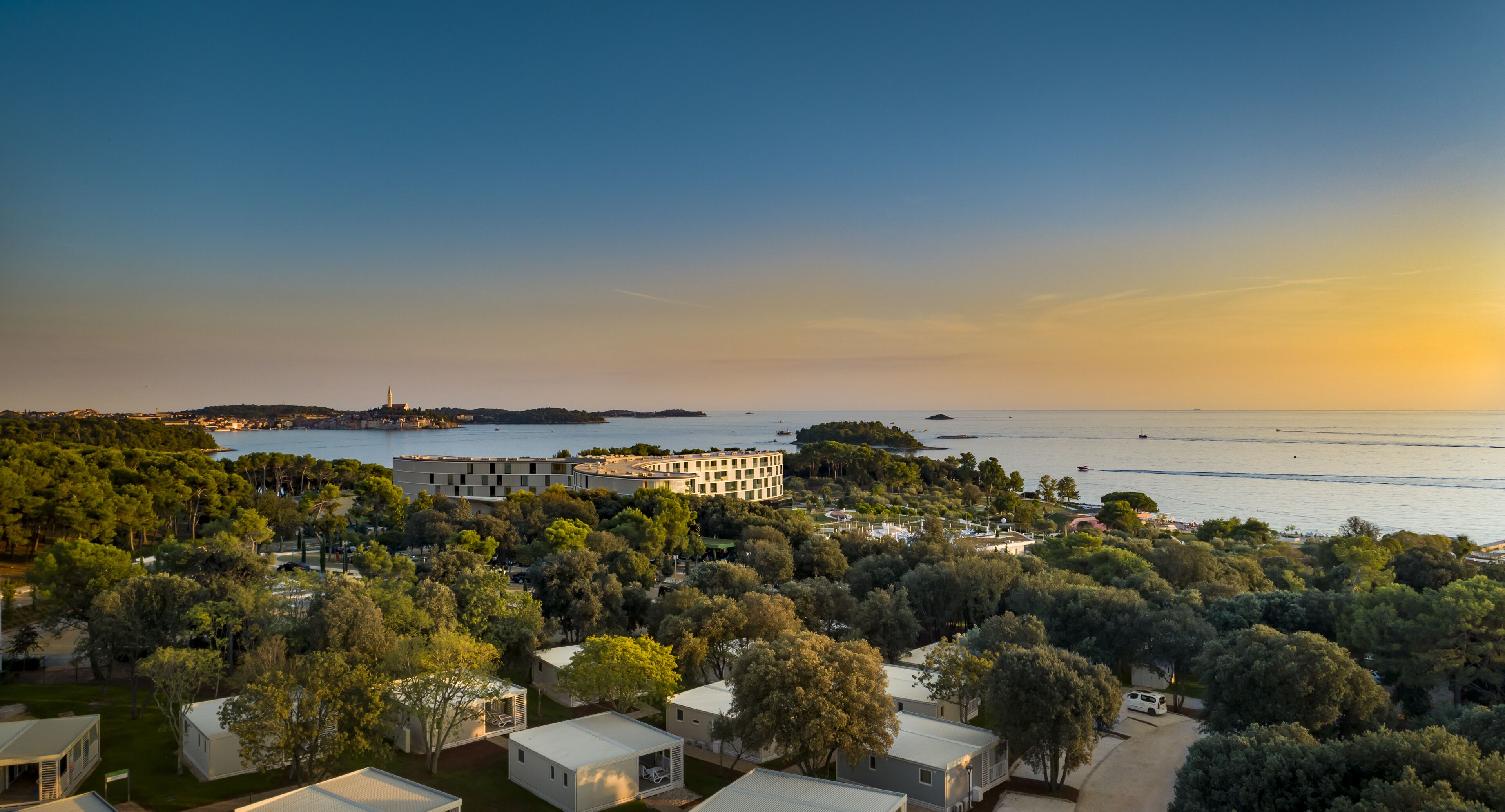 A coastal area with multiple small buildings surrounded by trees, a large body of water, and a distant island.