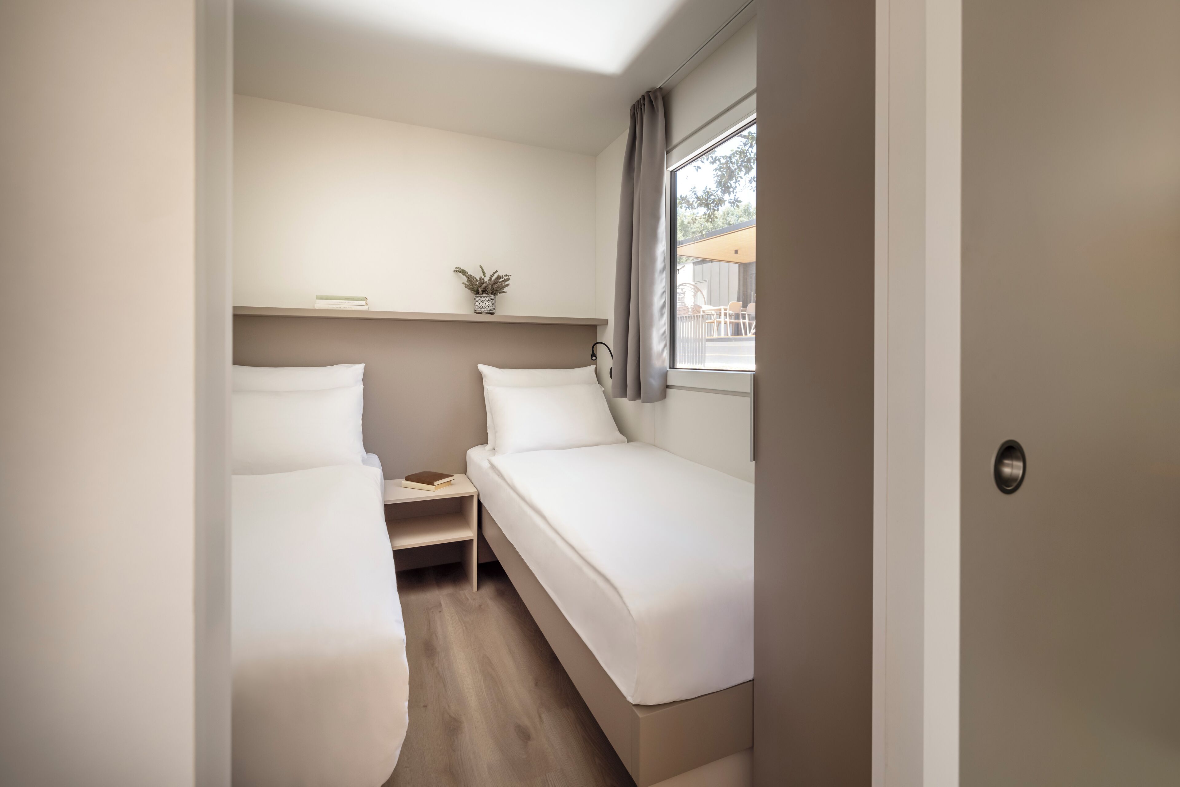 Une petite chambre moderne avec deux lits simples, une table de chevet, une étagère avec une plante et une fenêtre avec des rideaux.