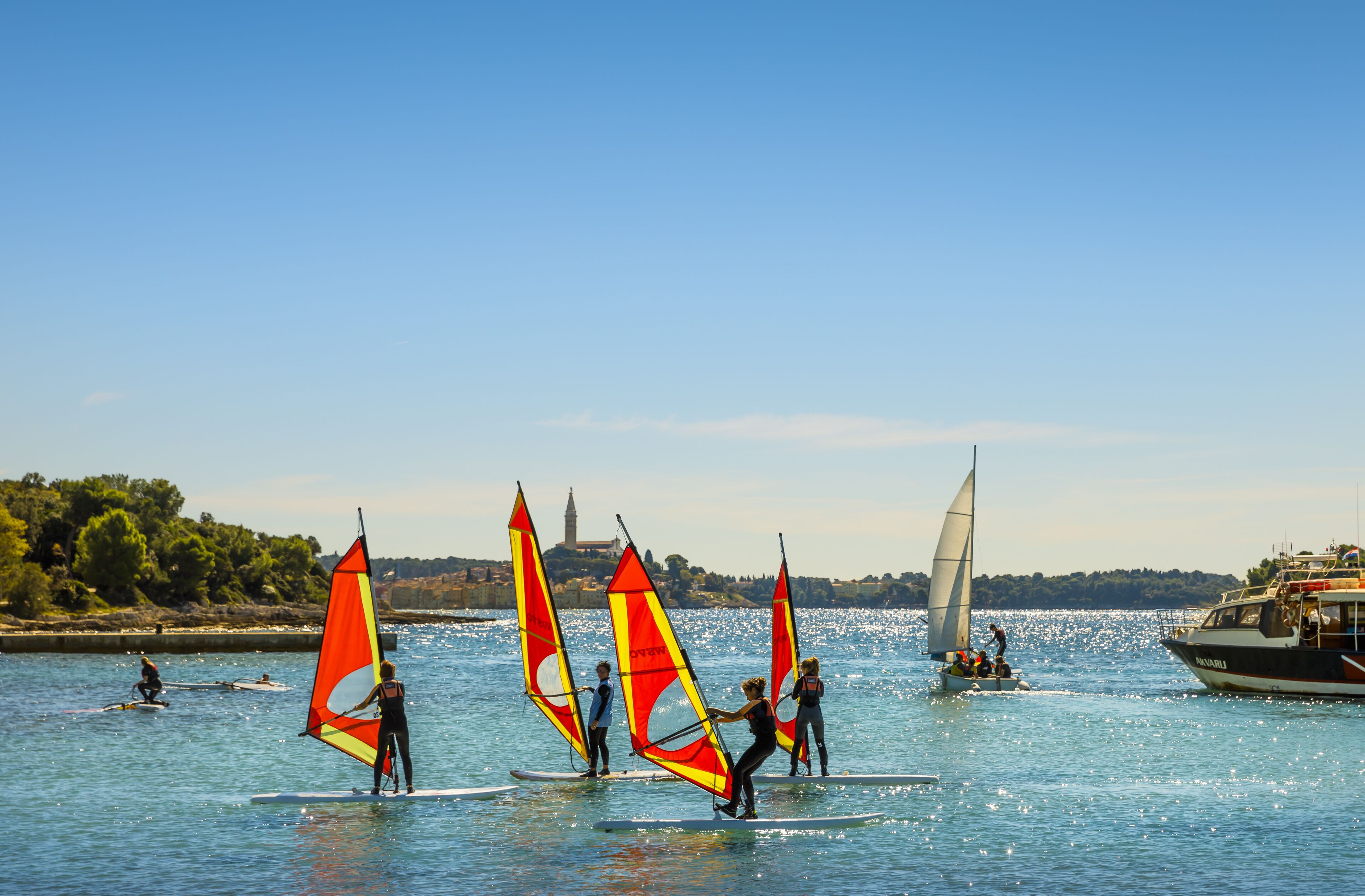 Un grupo de personas practicando windsurf en aguas tranquilas, con un barco al fondo.