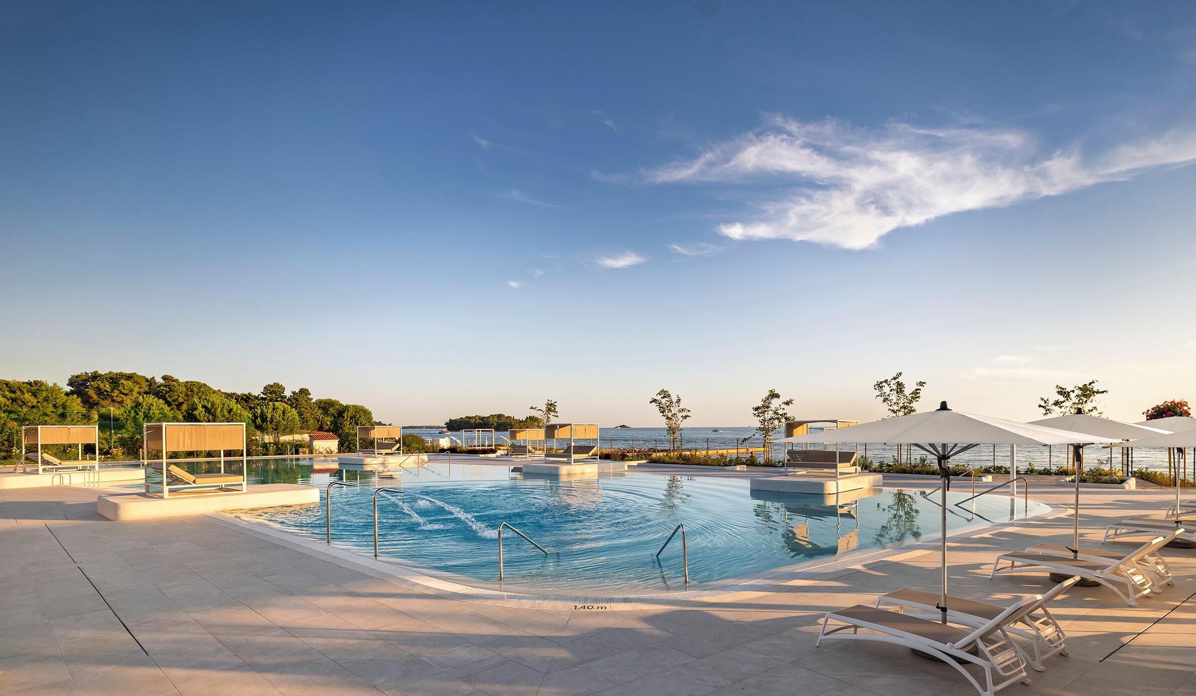 Ein großer Pool im Freien mit Liegestühlen und Sonnenschirmen, umgeben von Bäumen und Blick auf das Meer.